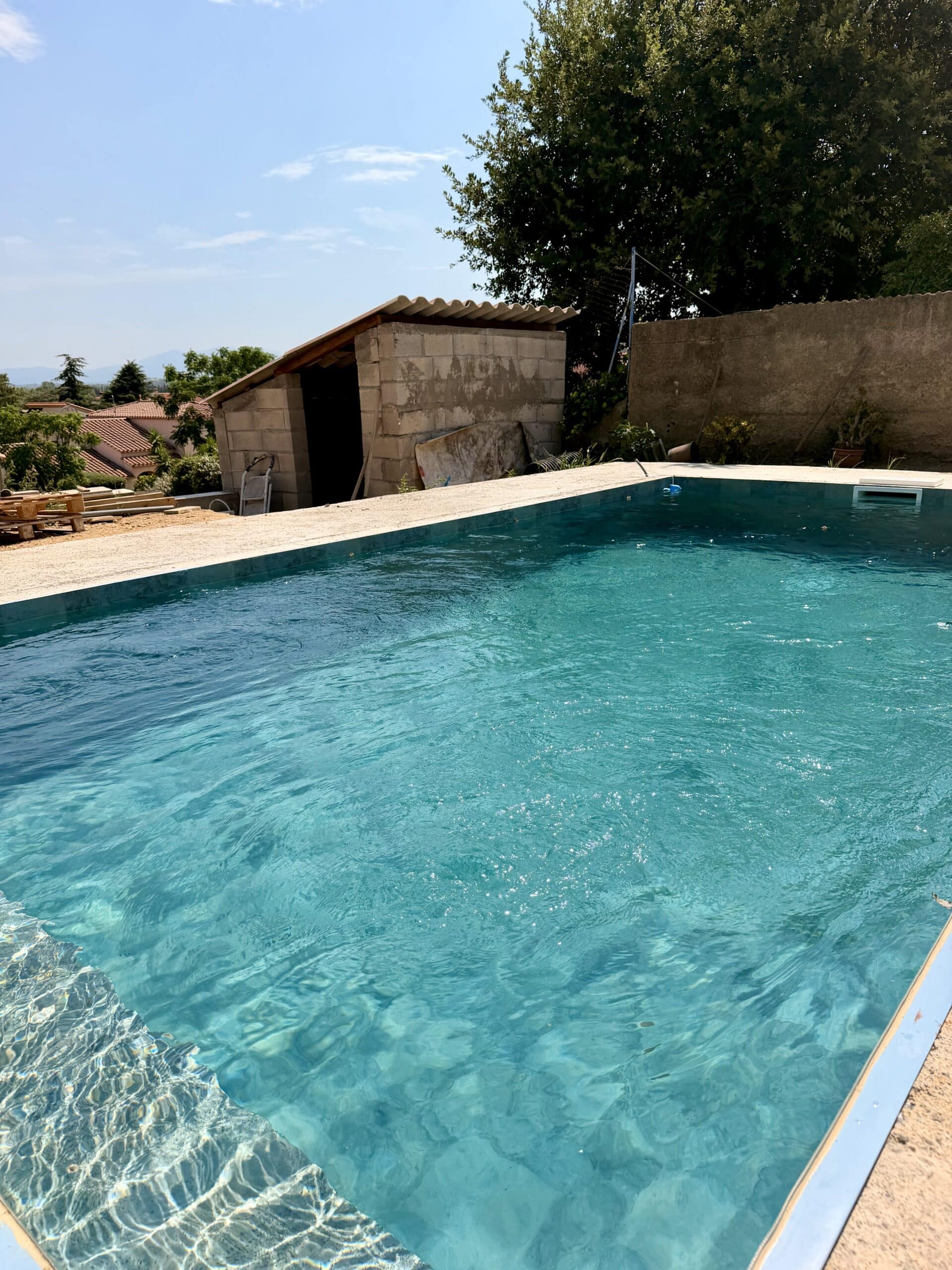 Piscine rectangulaire à l'eau turquoise avec un abri en parpaings et jardin verdoyant sous un ciel bleu.