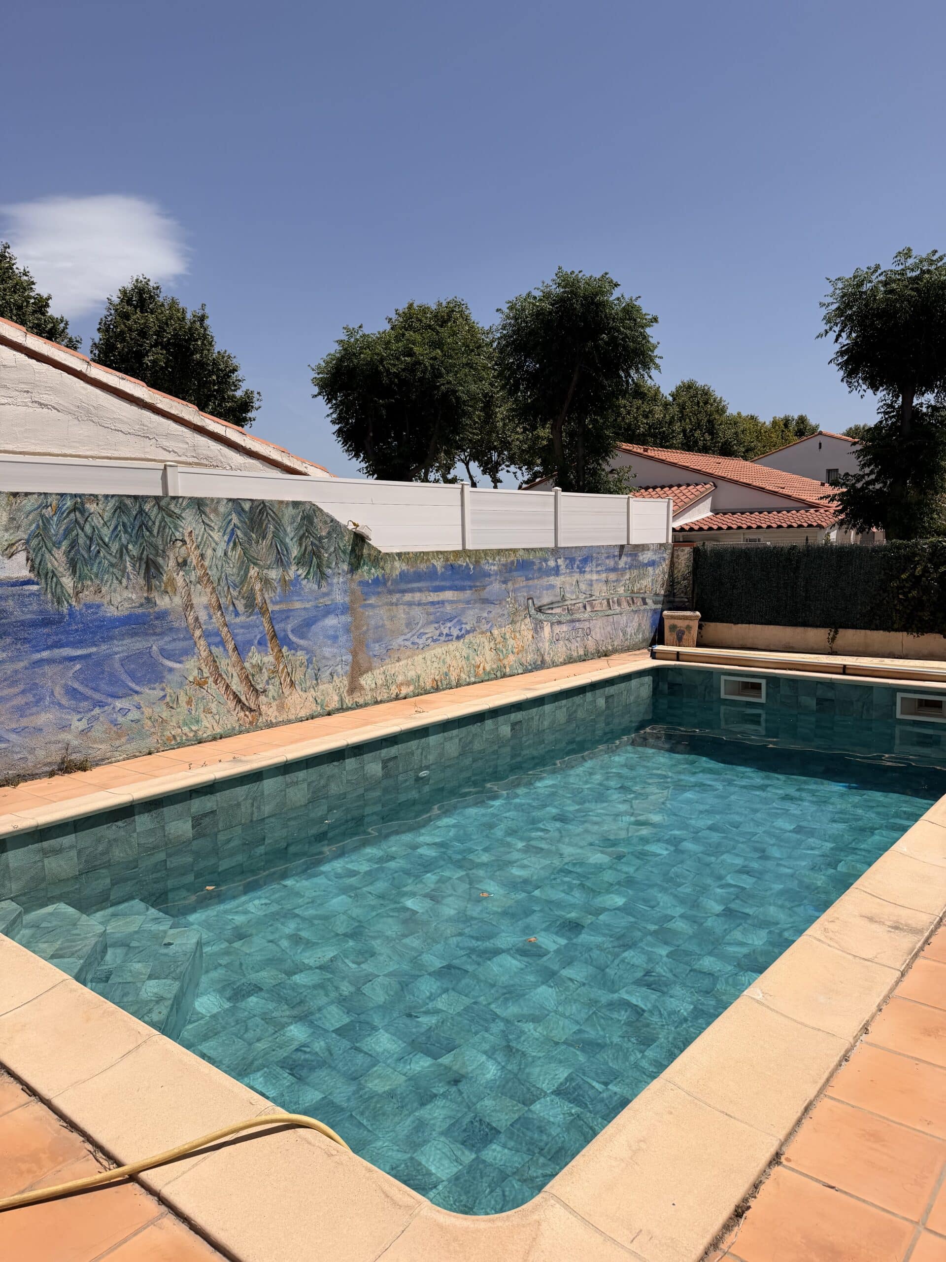 Piscine carrelée turquoise devant une fresque murale de plage et de palmiers, sous un grand ciel bleu d'été.