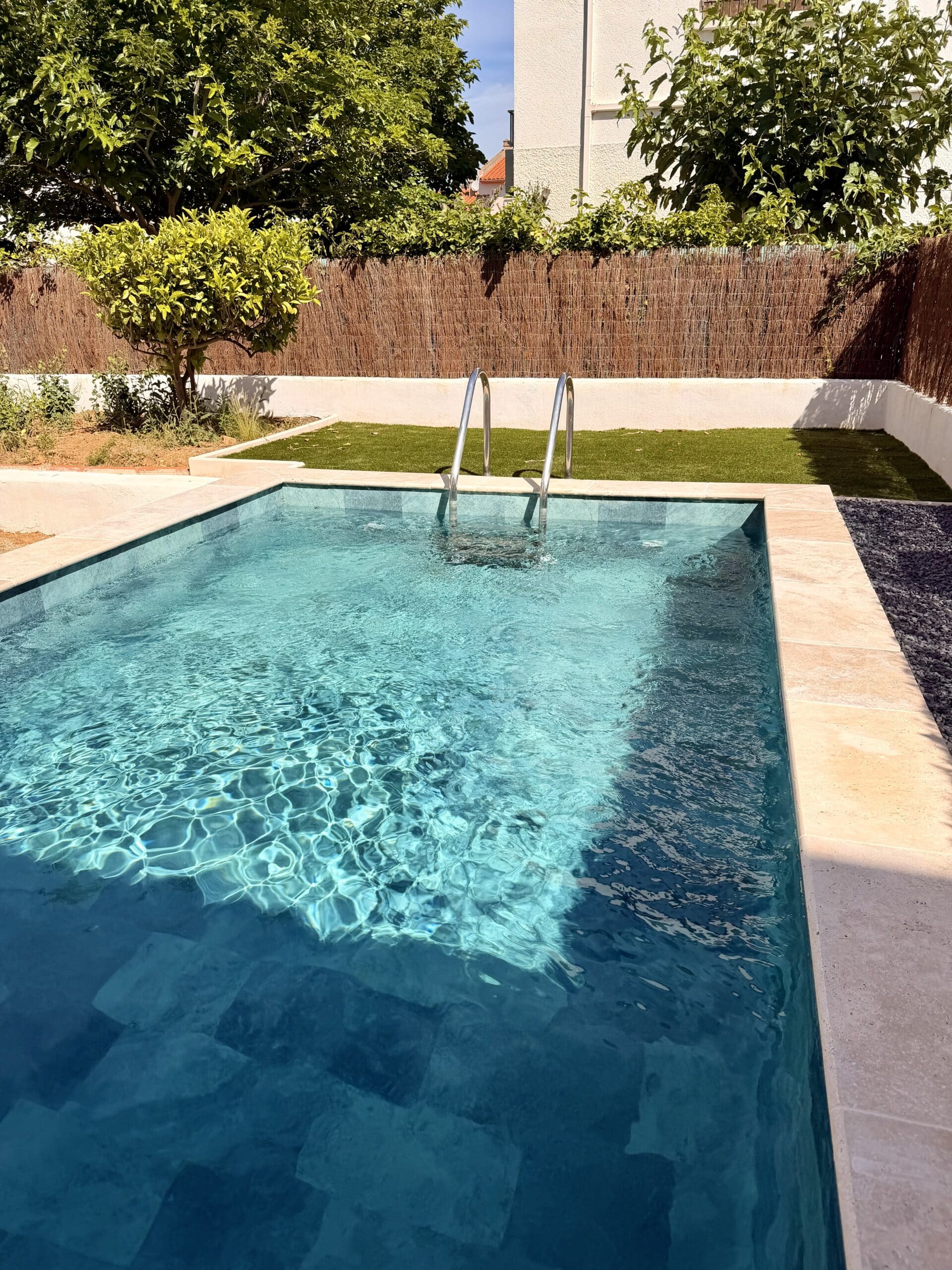 Piscine rectangulaire à l'eau turquoise avec margelles en pierre beige et jardin verdoyant ensoleillé.