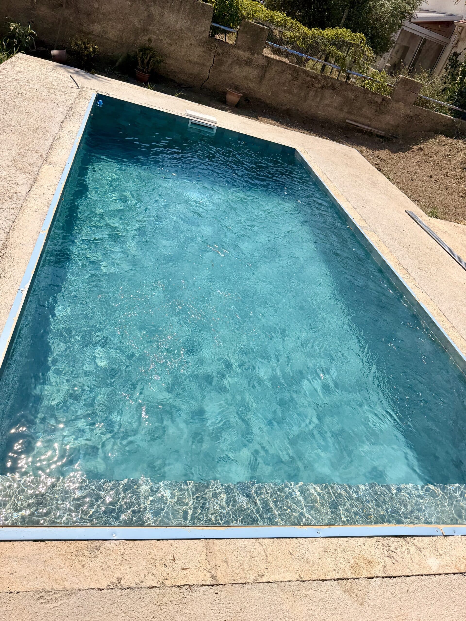 Piscine rectangulaire à l'eau turquoise cristalline sur une terrasse en béton ensoleillée avec mur en pierre.