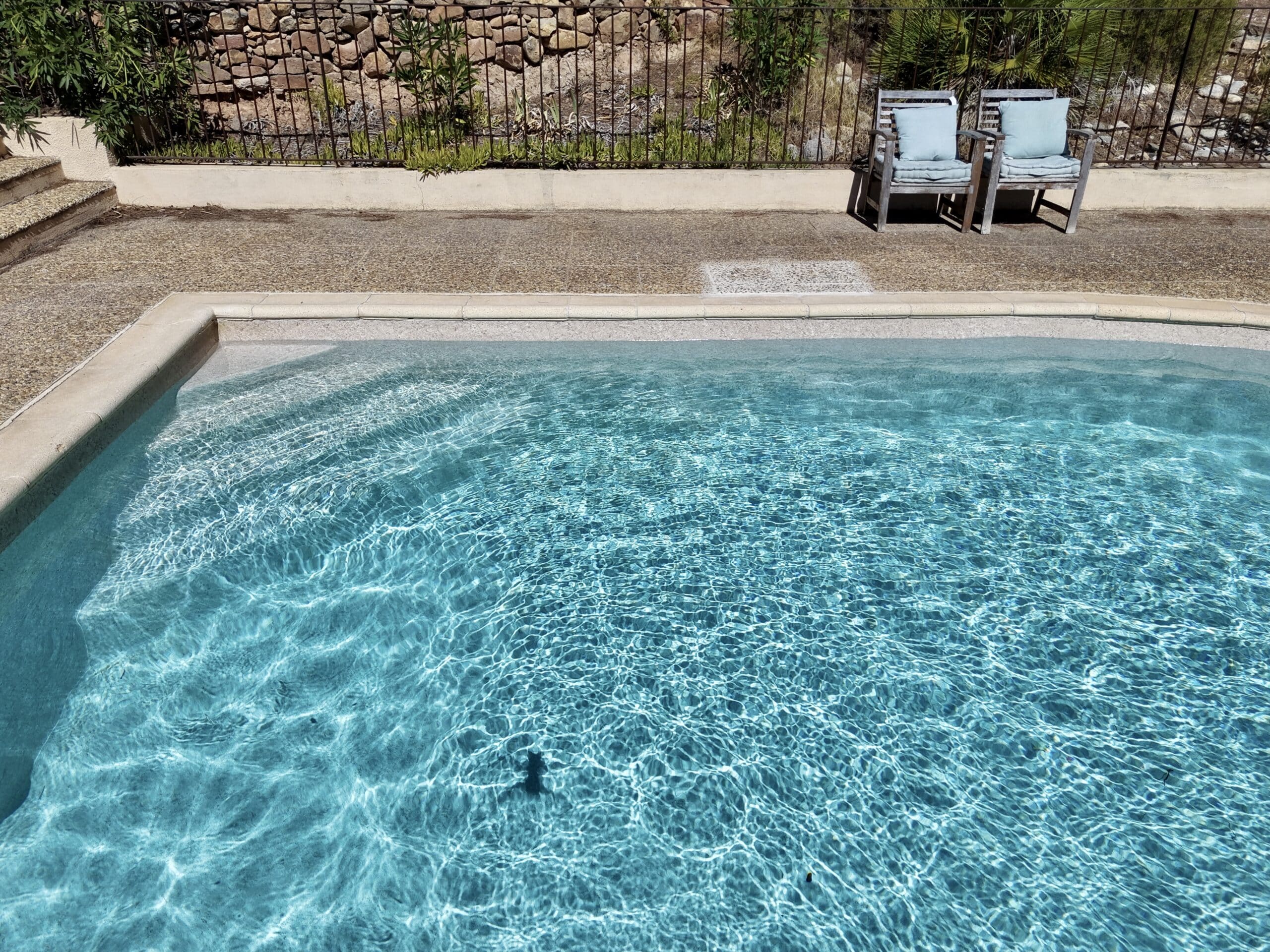 Piscine à l'eau turquoise scintillante, terrasse avec deux fauteuils bleus et mur de pierres en arrière-plan.
