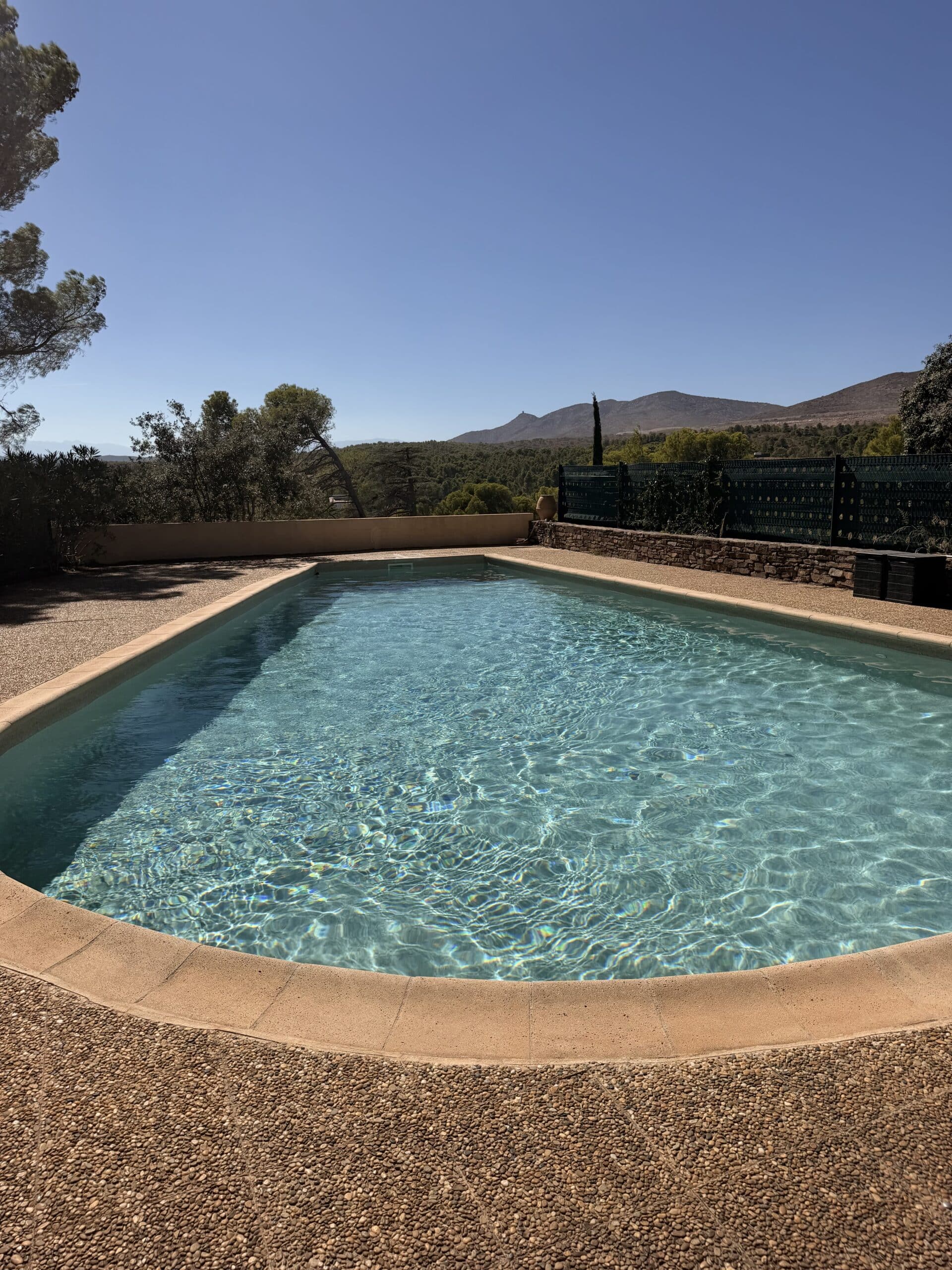 Piscine extérieure à l'eau turquoise avec bordure en pierre, vue sur des collines boisées sous un ciel bleu dégagé.