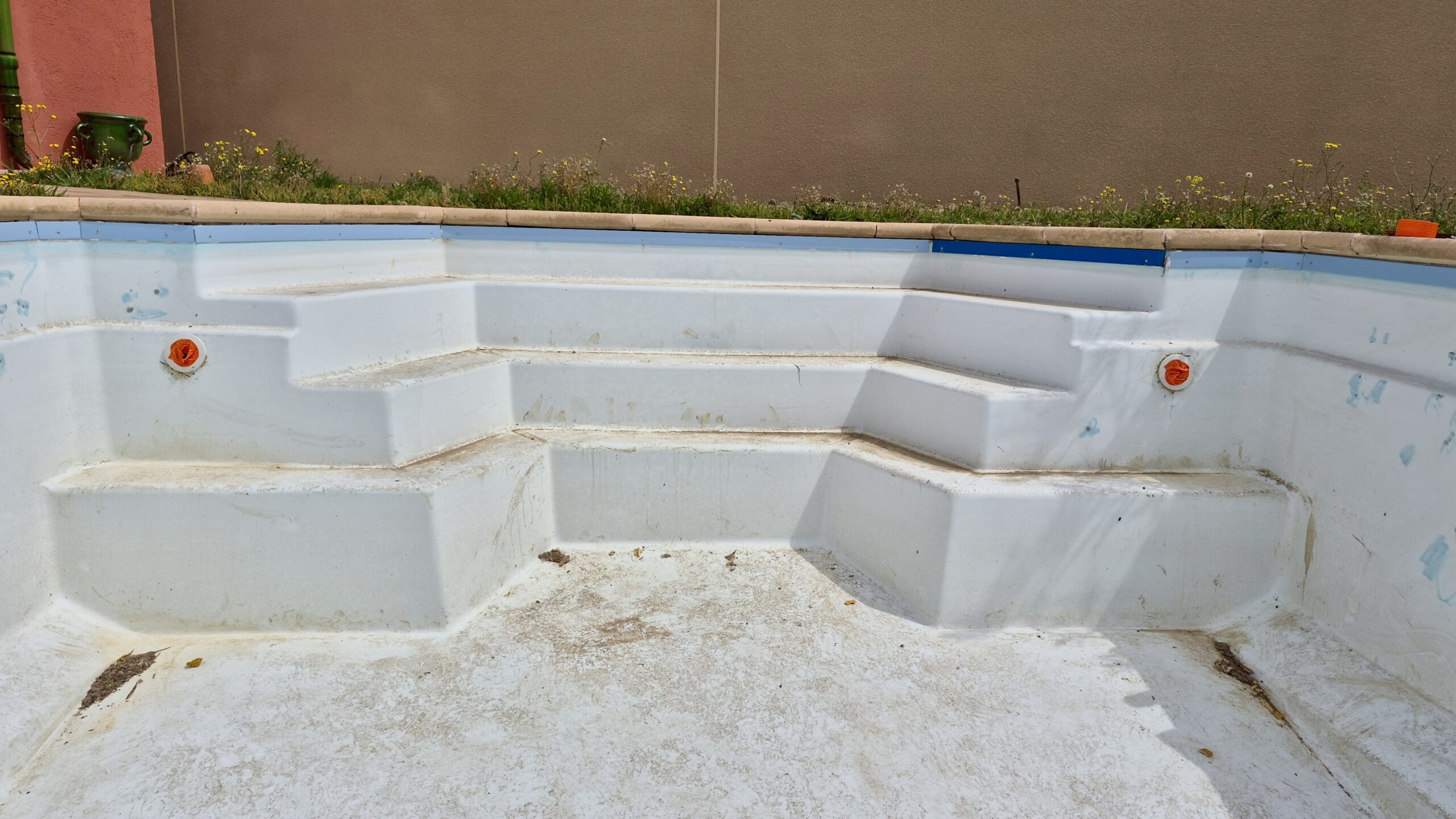 Escalier d'une piscine vide en coque blanche montrant des signes d'usure et des buses bouchées avant rénovation.