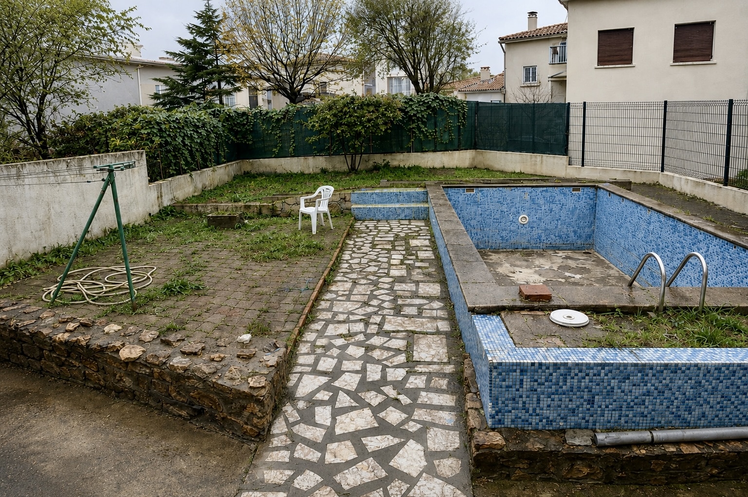Piscine abandonnée carrelée de bleu dans un jardin en friche avec dallage en pierre et chaise blanche.