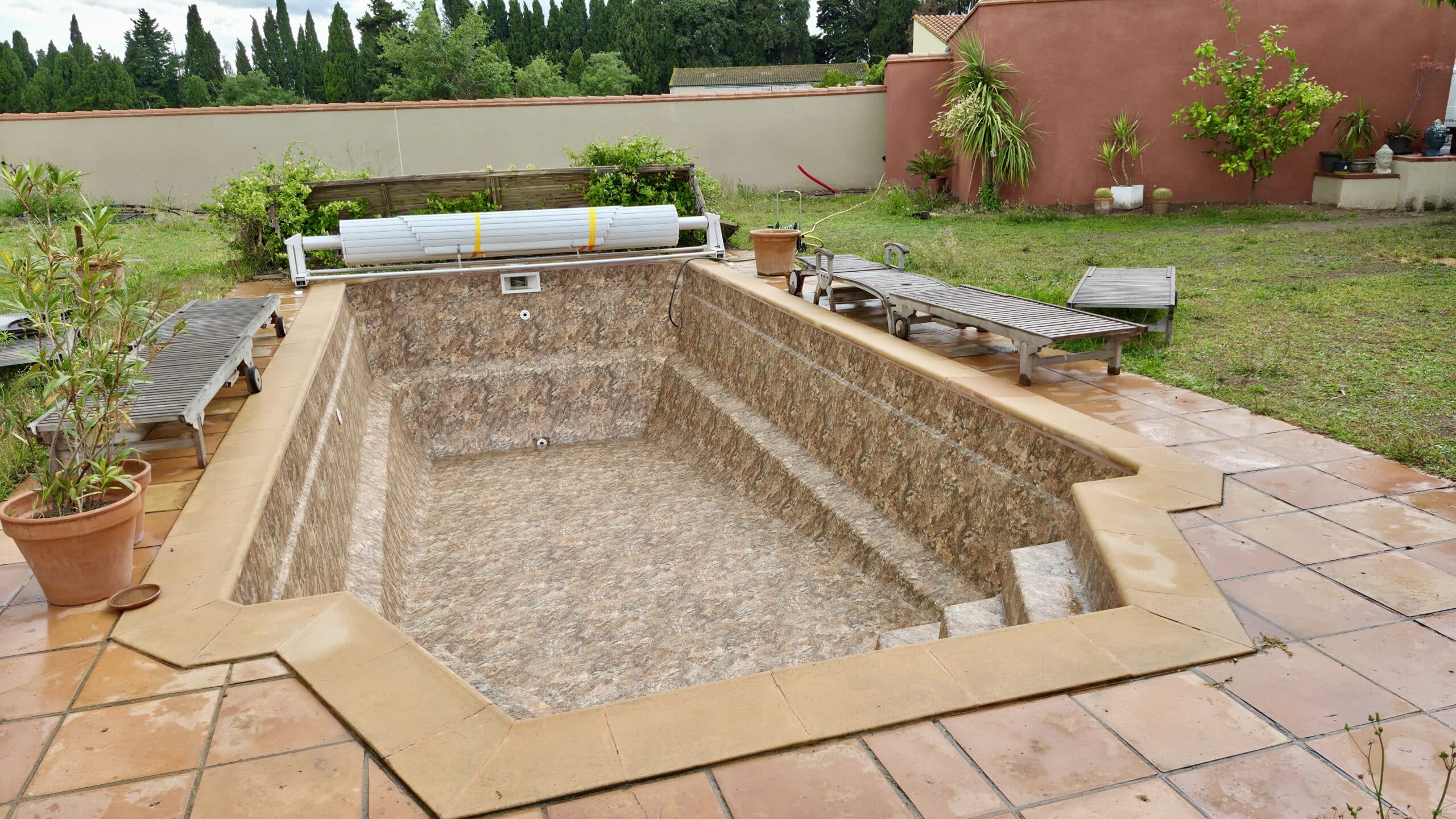 Piscine vide à liner marbré beige entourée d'une terrasse en terre cuite et de chaises longues en bois.
