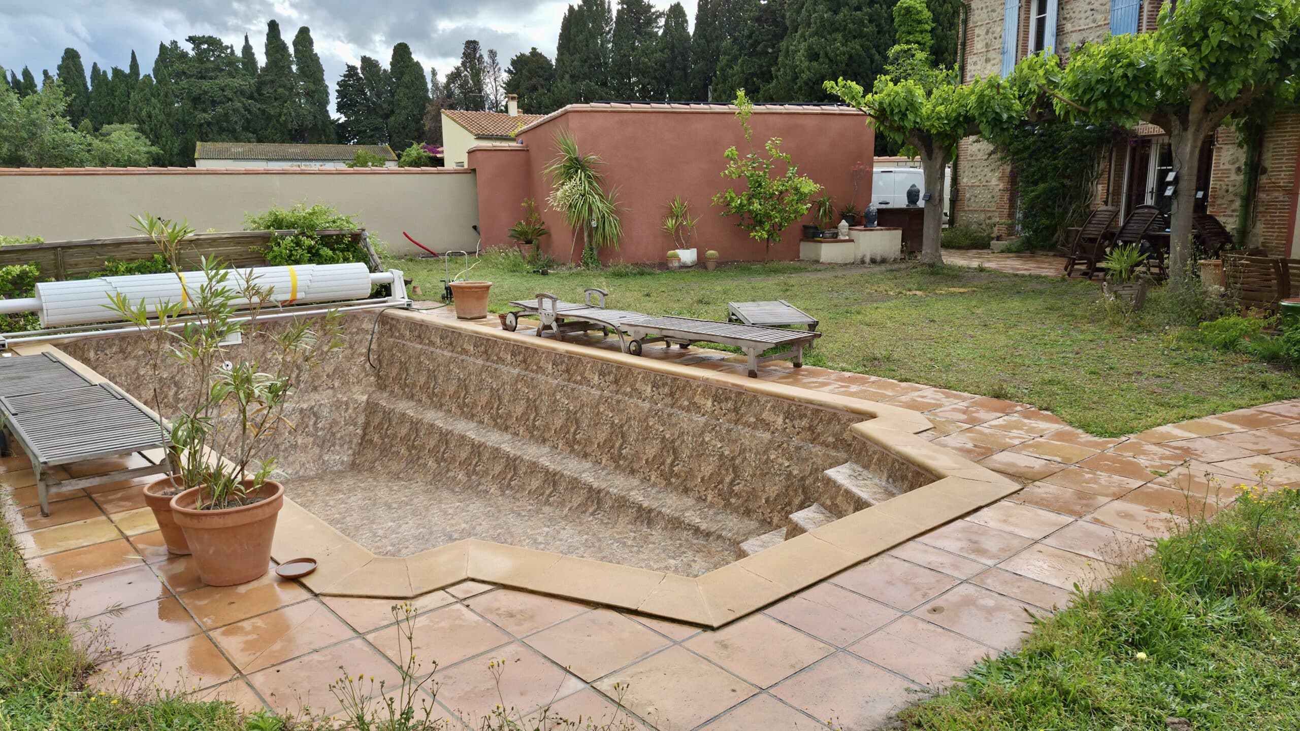 Piscine vide en pierre avec terrasse en carrelage, chaises longues en bois et jardin verdoyant sous un ciel nuageux.