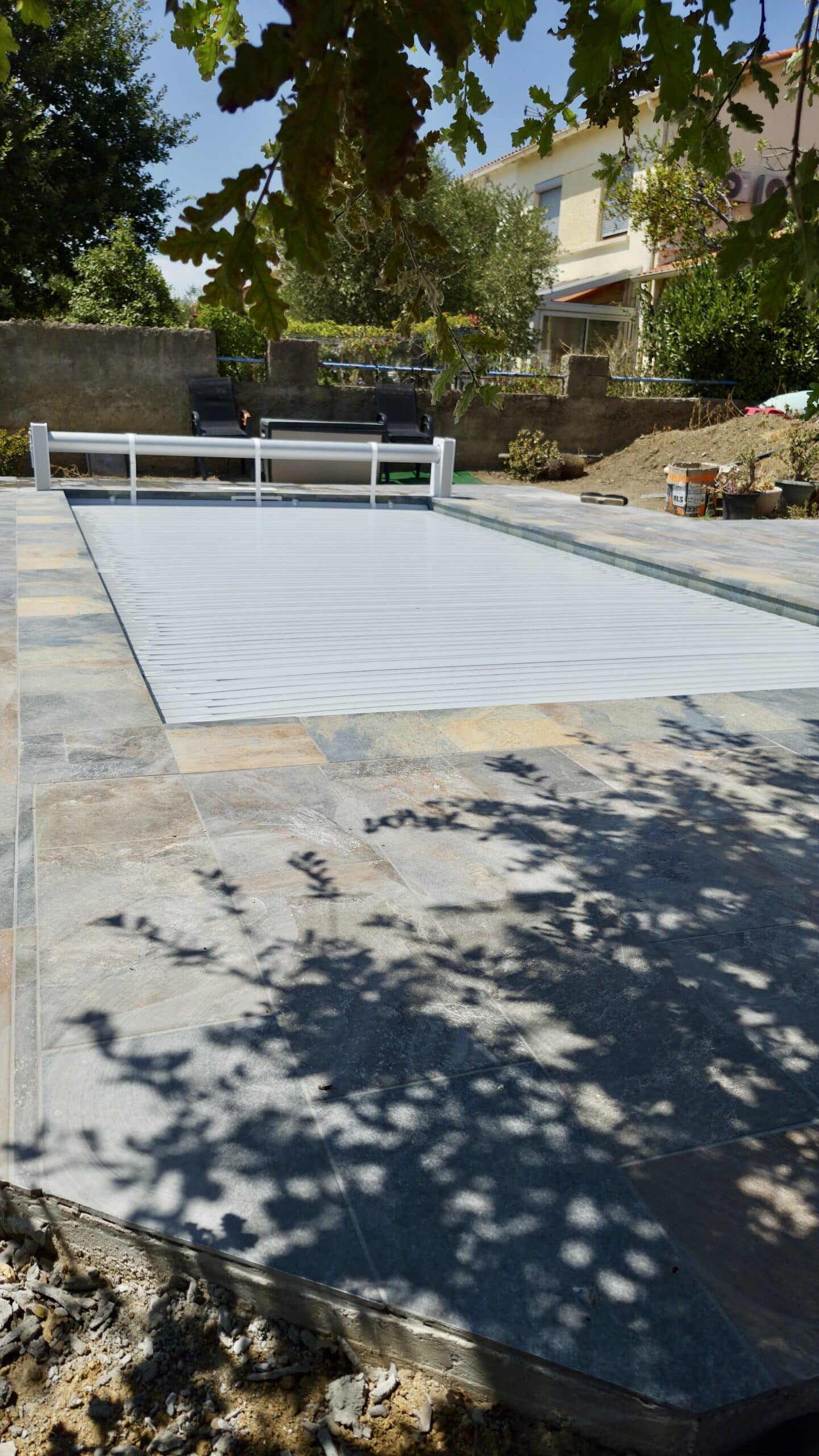 Piscine avec volet roulant automatique gris sur une terrasse en pierre naturelle ombragée par des arbres.