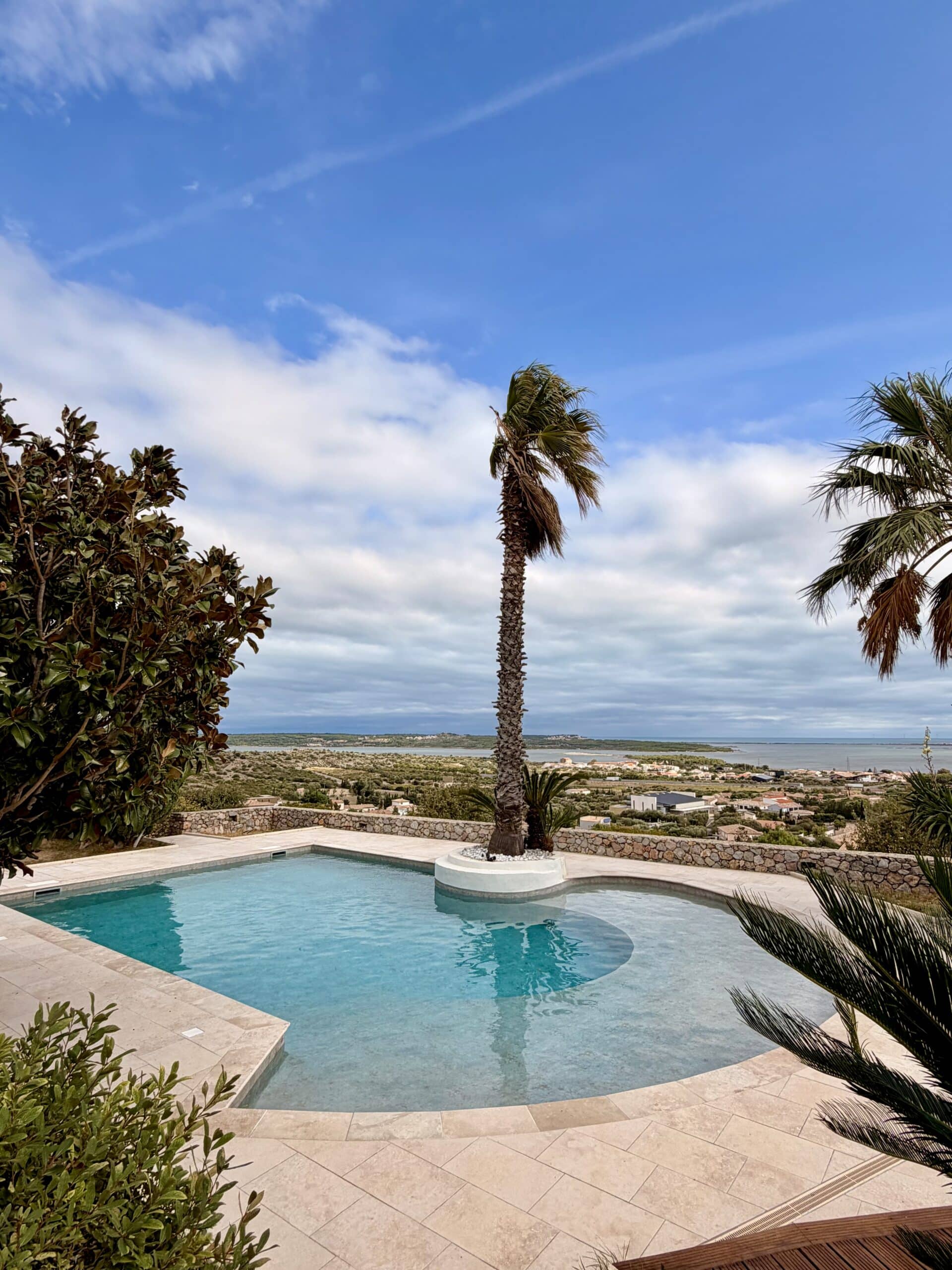 Piscine luxueuse avec palmier central offrant une vue panoramique sur la mer et un paysage méditerranéen ensoleillé.