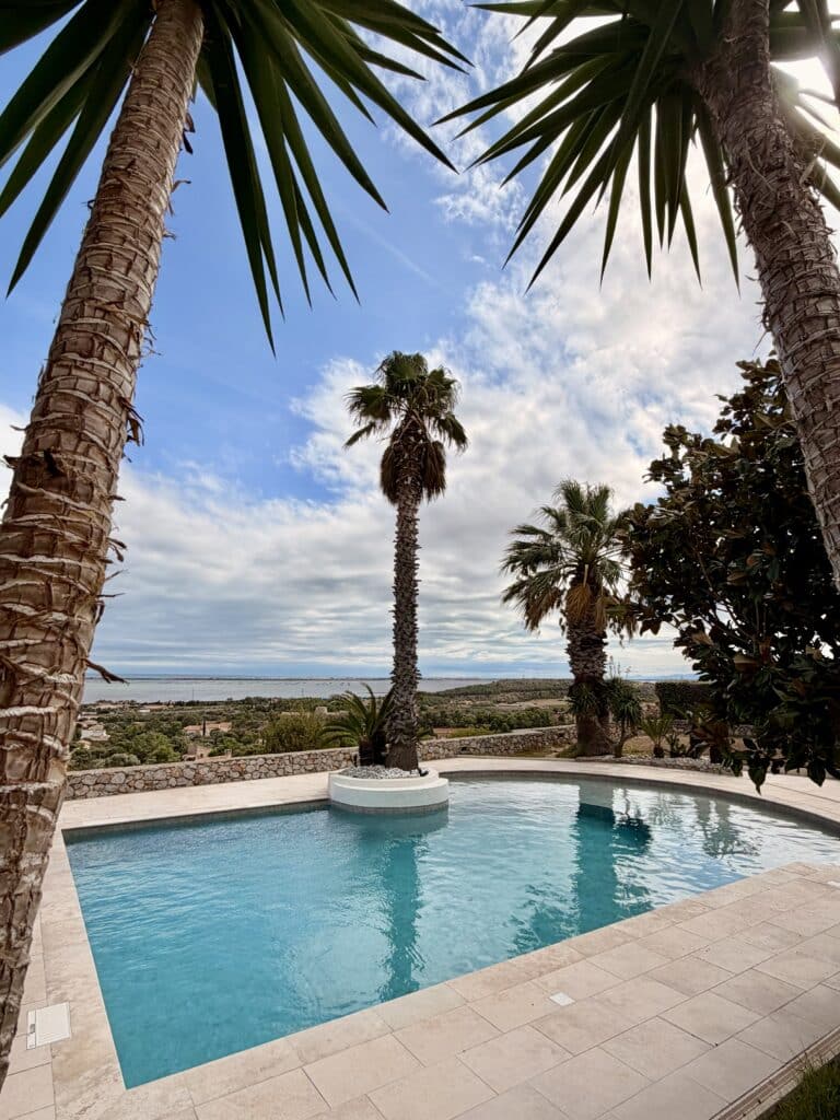 Piscine avec vue mer et palmiers dans un jardin de luxe Piscine azur bordée de palmiers offrant une vue panoramique sur la mer et la côte sous un ciel ensoleillé.