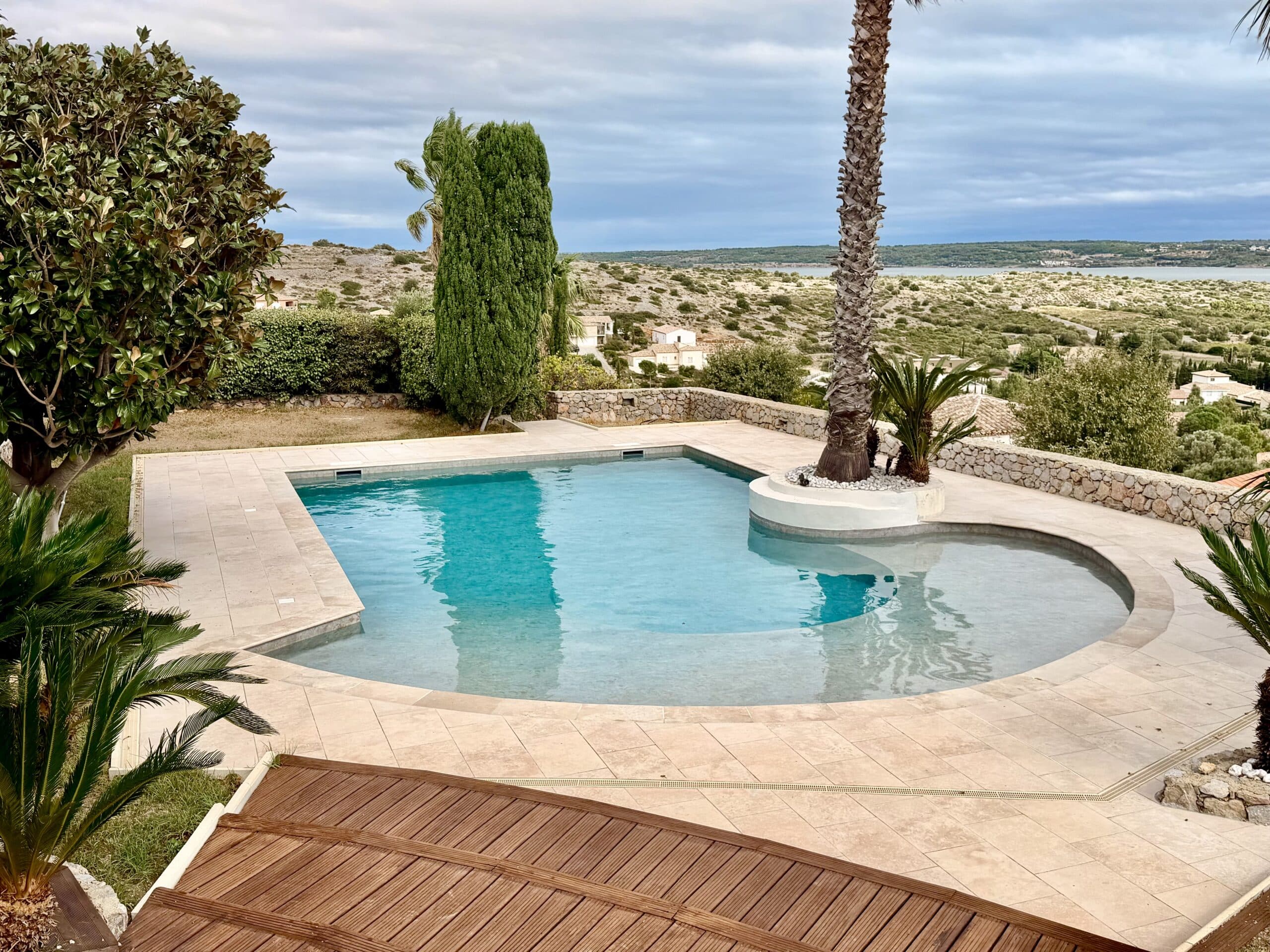 Piscine moderne avec terrasse en pierre, palmier et vue panoramique sur la mer et le paysage vallonné.