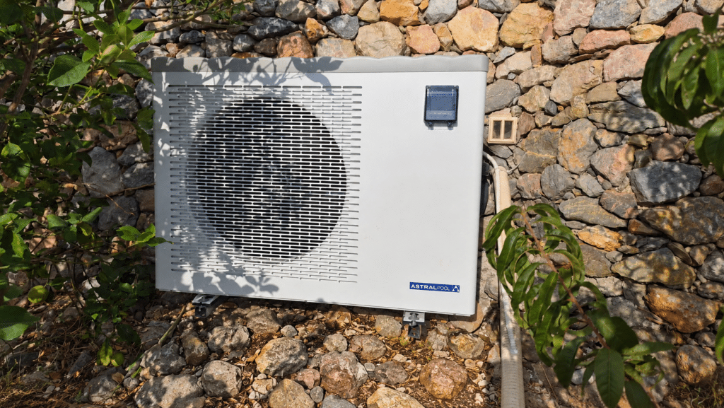 Pompe à chaleur AstralPool : installation piscine efficace Pompe à chaleur de piscine AstralPool installée devant un mur en pierres sèches dans un jardin.