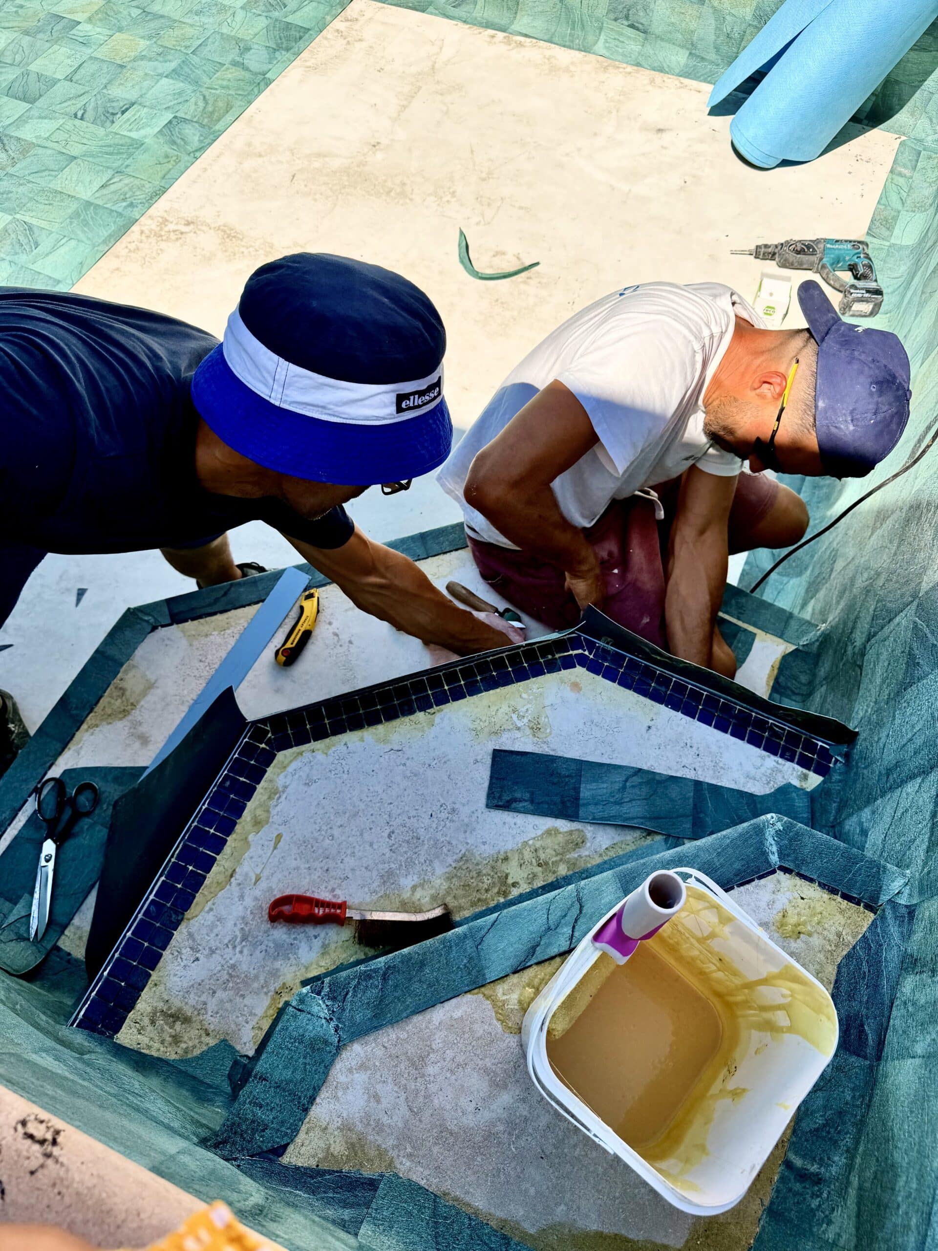 Deux ouvriers installent un liner de piscine vert texturé sur des marches avec colle, ciseaux et cutter de précision.
