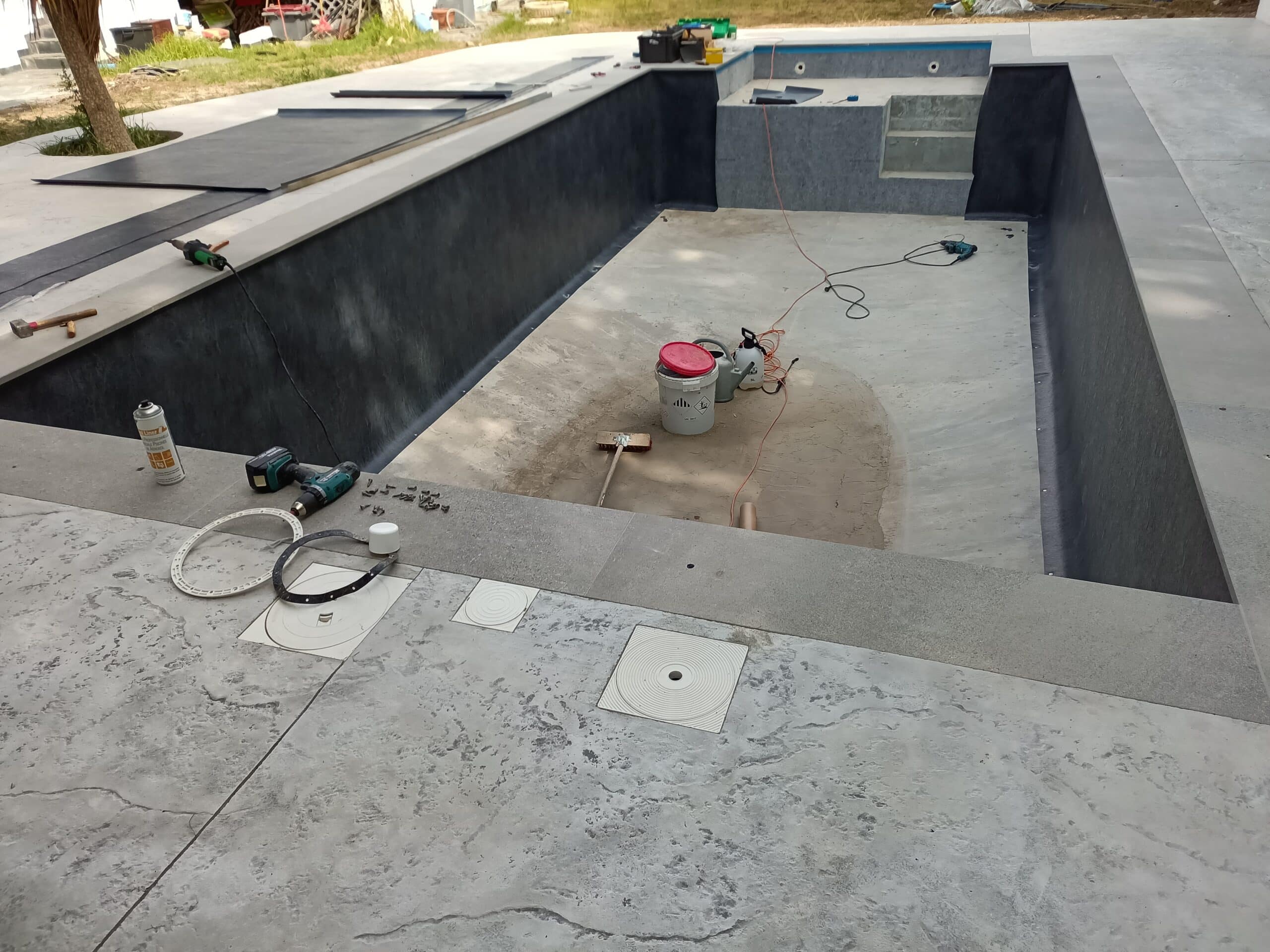 Chantier de piscine avec pose de liner gris anthracite, outils de construction et plage en béton décoratif.