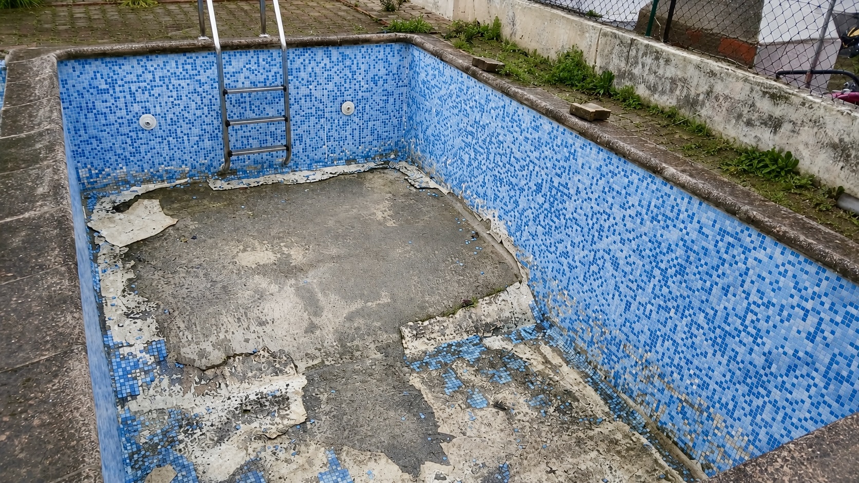 Piscine carrelée en mauvais état avec des mosaïques bleues qui se décollent du fond en béton et échelle en métal.