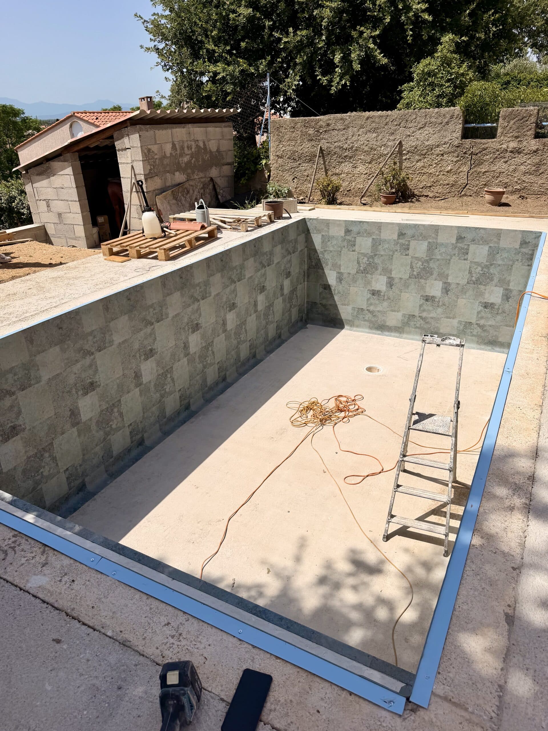 Piscine en rénovation avec murs carrelés, échelle intérieure et bordure bleue, chantier extérieur sous un ciel dégagé.