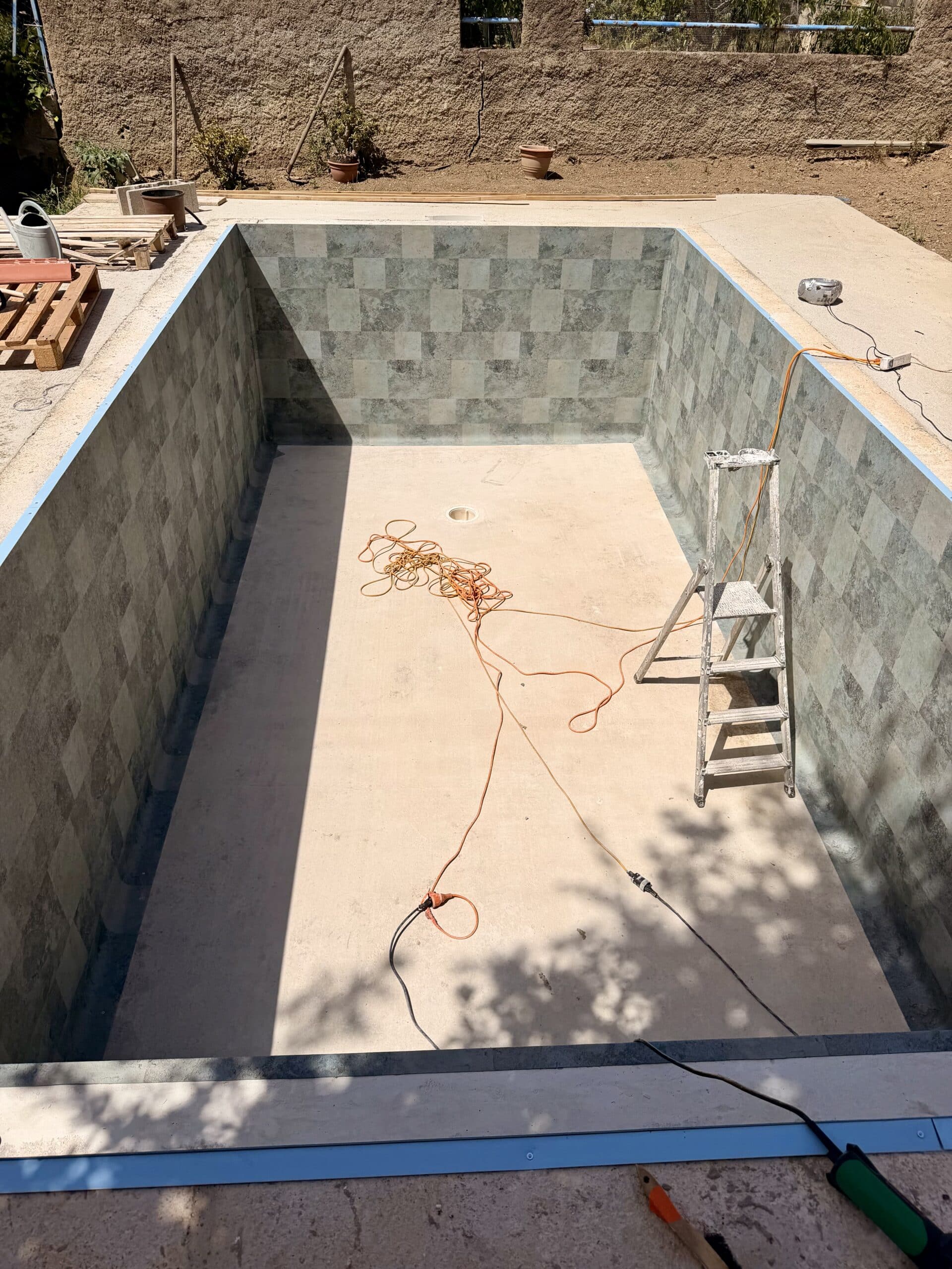 Chantier de rénovation d'une piscine vide avec carrelage gris, échelle et rallonge électrique au soleil.