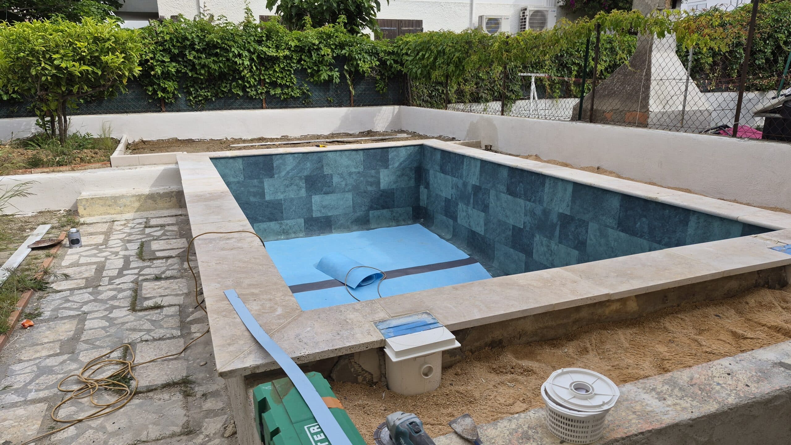 Chantier d'une piscine en rénovation avec carrelage bleu canard, margelles en travertin et liner en cours de pose.