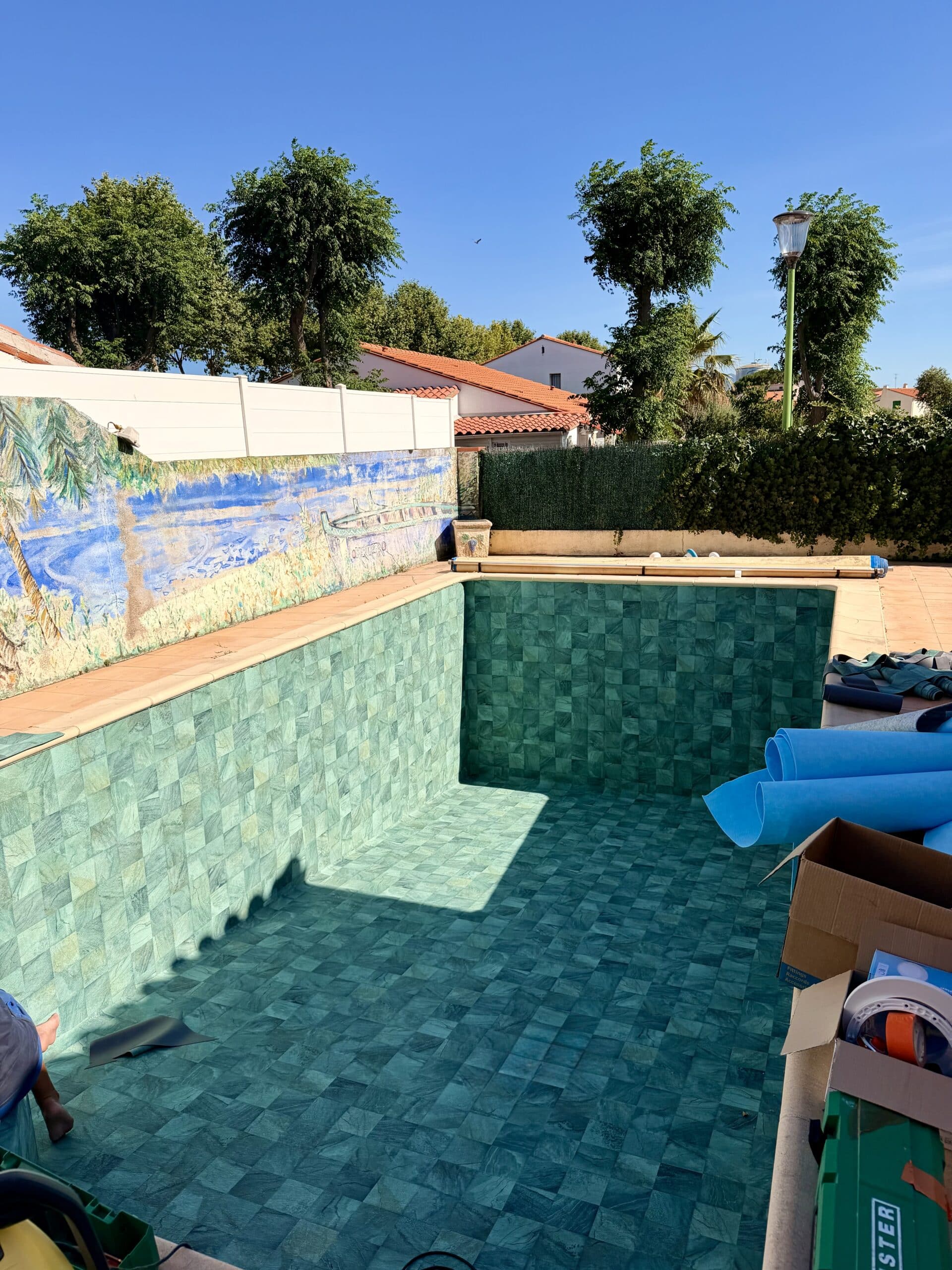Rénovation de piscine : bassin vide avec mosaïque verte, fresque murale et matériel de pose sous un ciel bleu.
