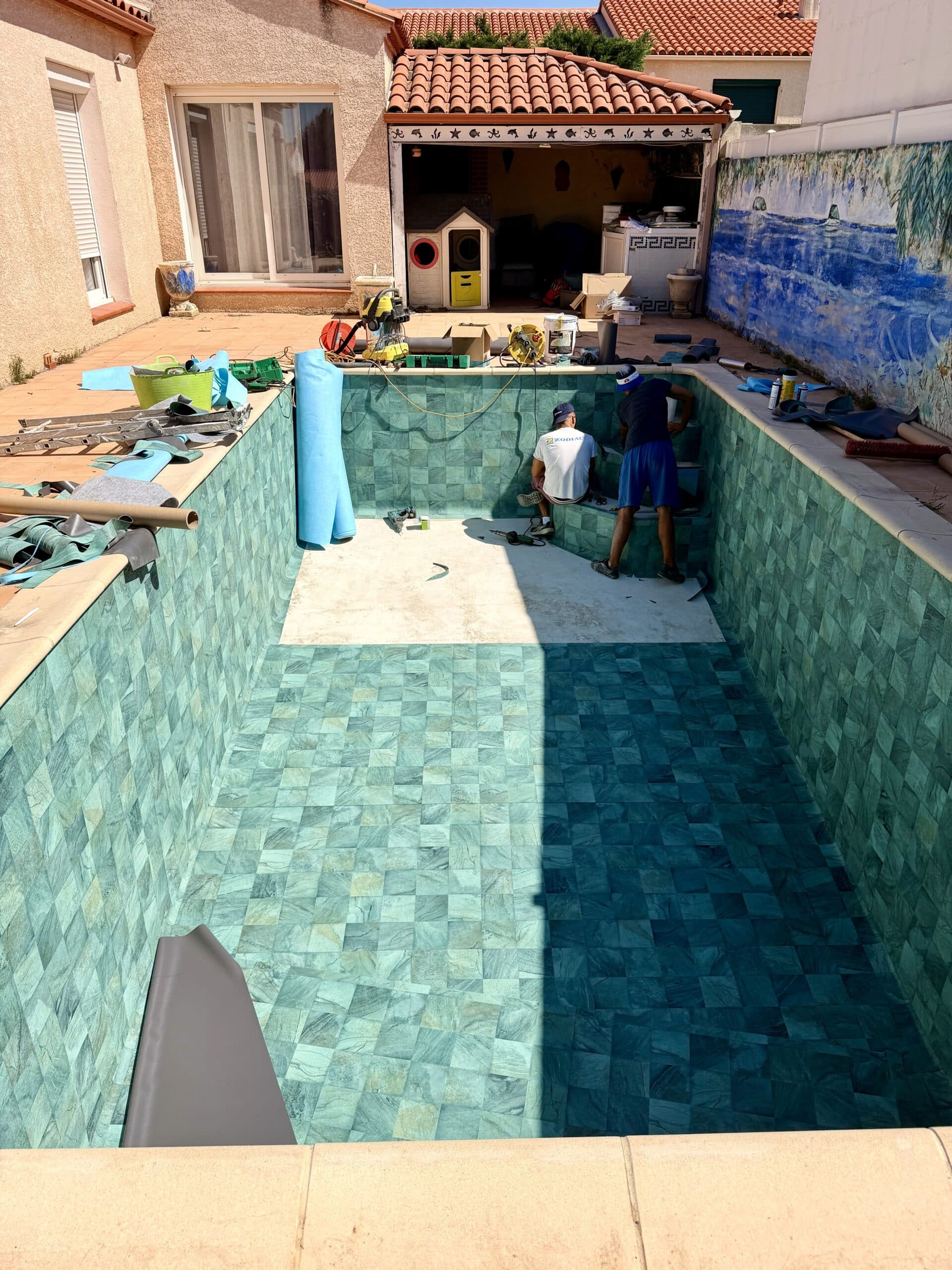 Deux ouvriers rénovent une piscine avec des carreaux effet pierre verte, outils et liner à disposition sur le rebord.