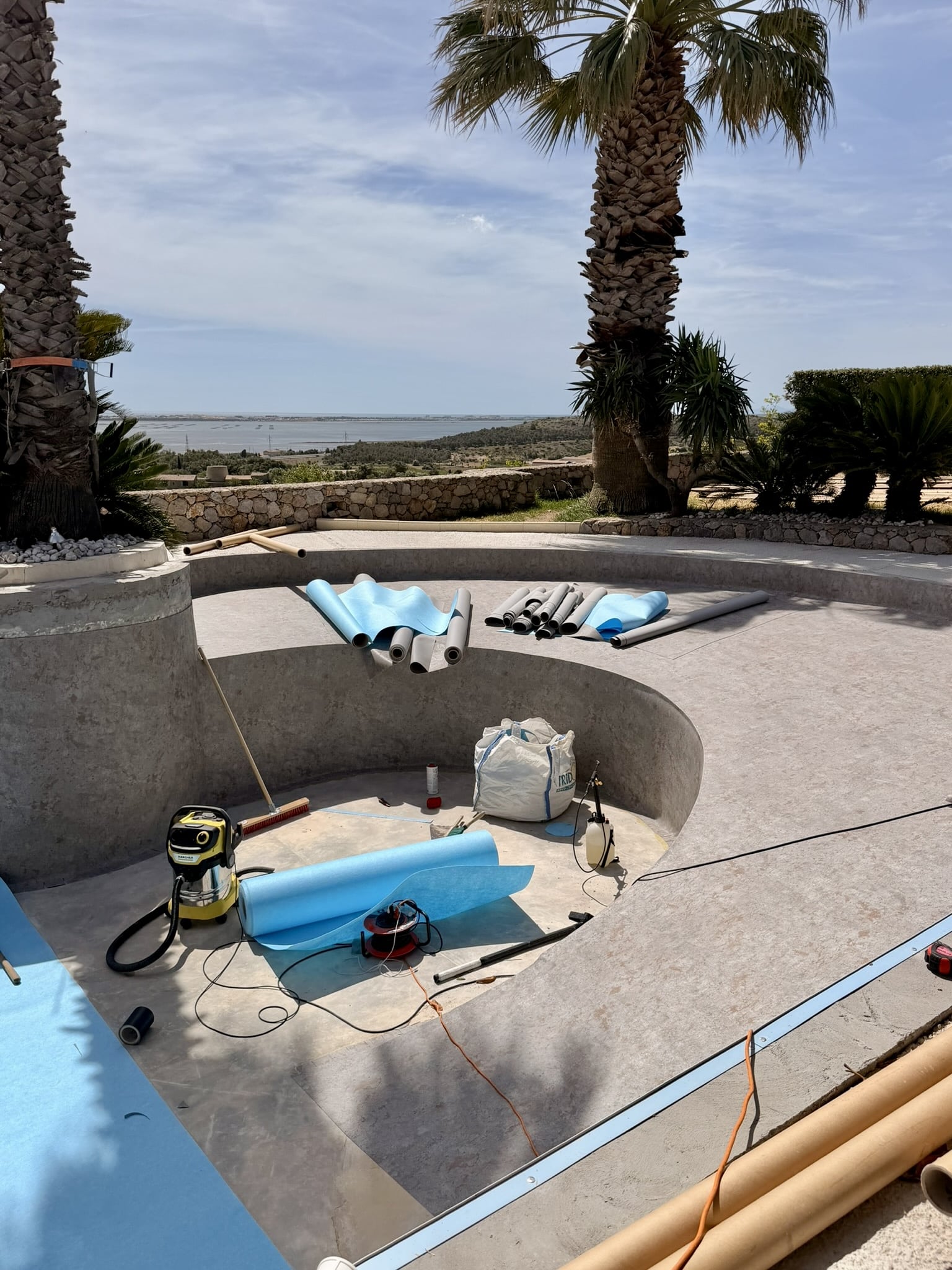 Rénovation de piscine avec pose de liner bleu, outillage Karcher et vue mer panoramique sous les palmiers.