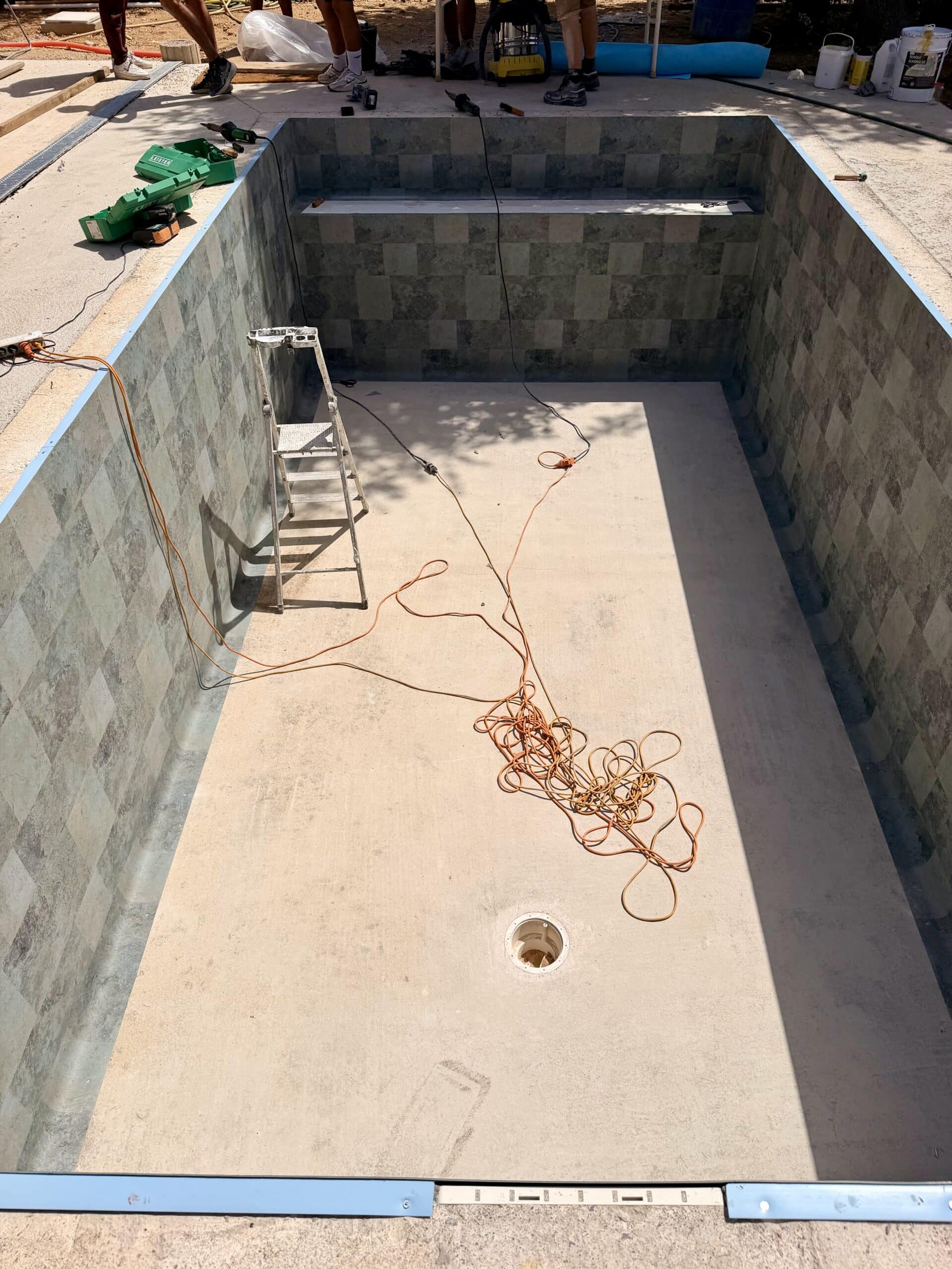 Chantier de piscine avec liner gris à motifs, échelle et câbles sur le fond beige en plein soleil.