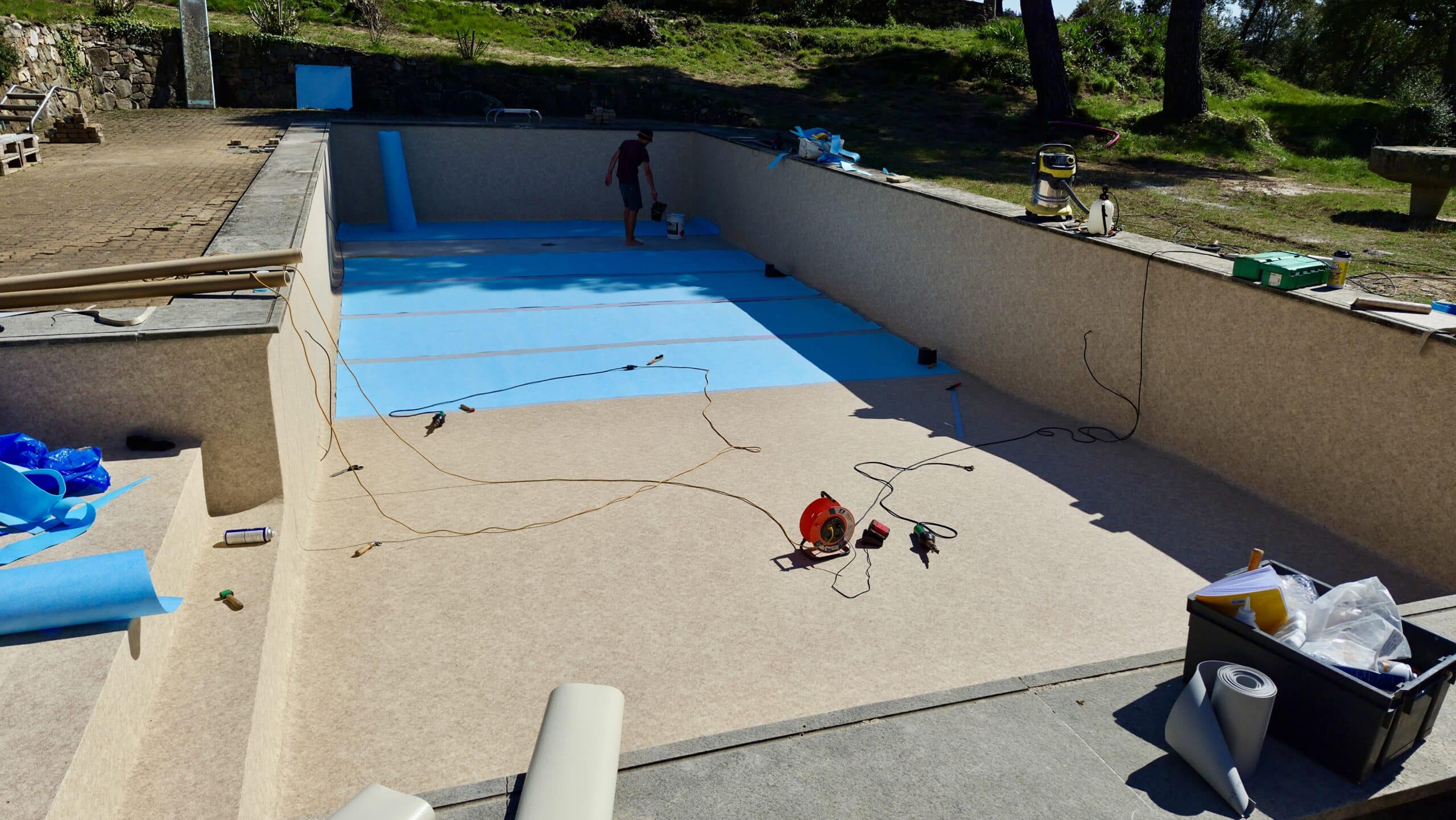 Pose d'un liner bleu dans une piscine en rénovation. Un technicien travaille sur le fond du bassin avec ses outils.