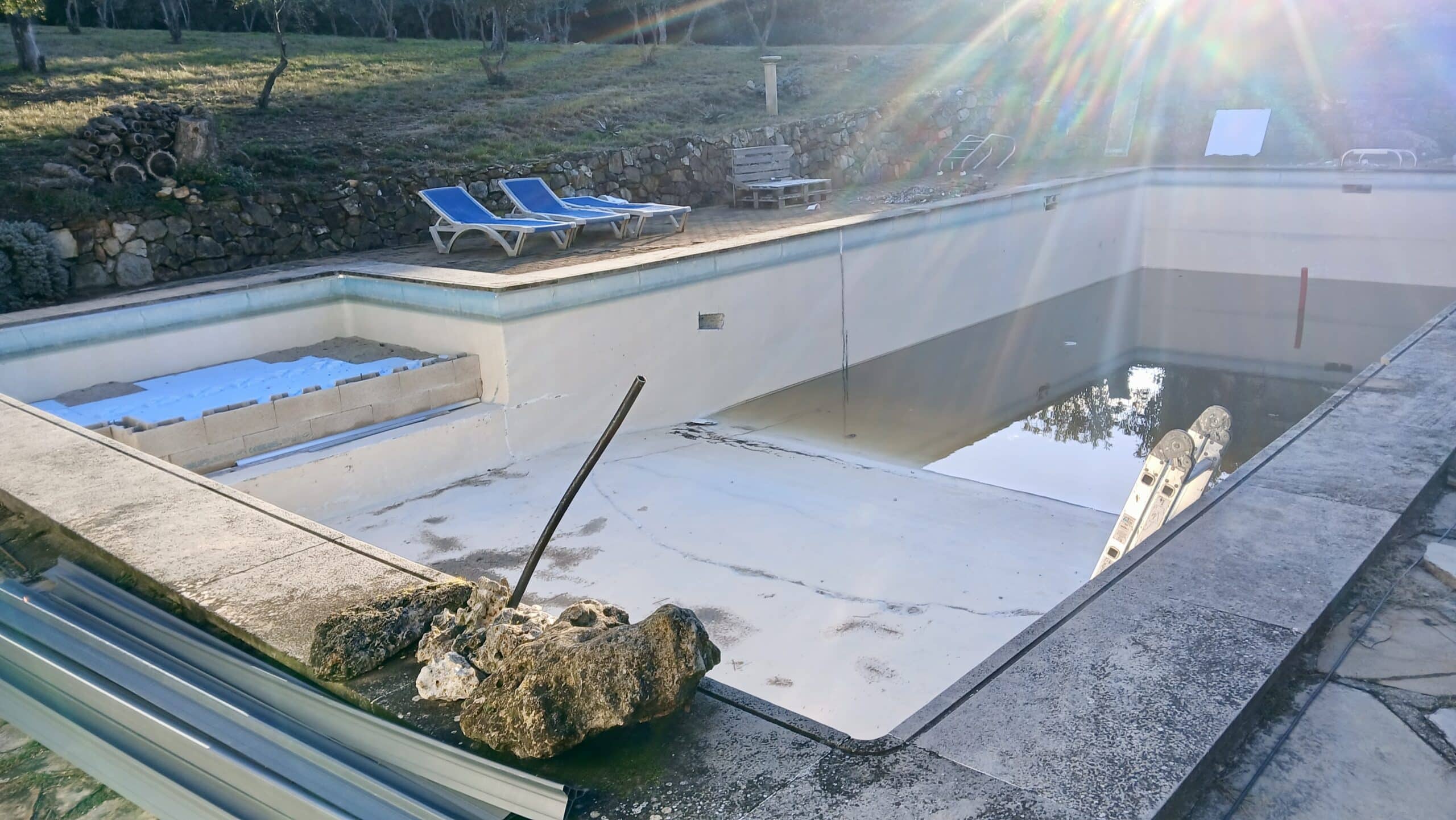 Piscine vide en rénovation avec transats bleus et muret en pierre sous un soleil éclatant.