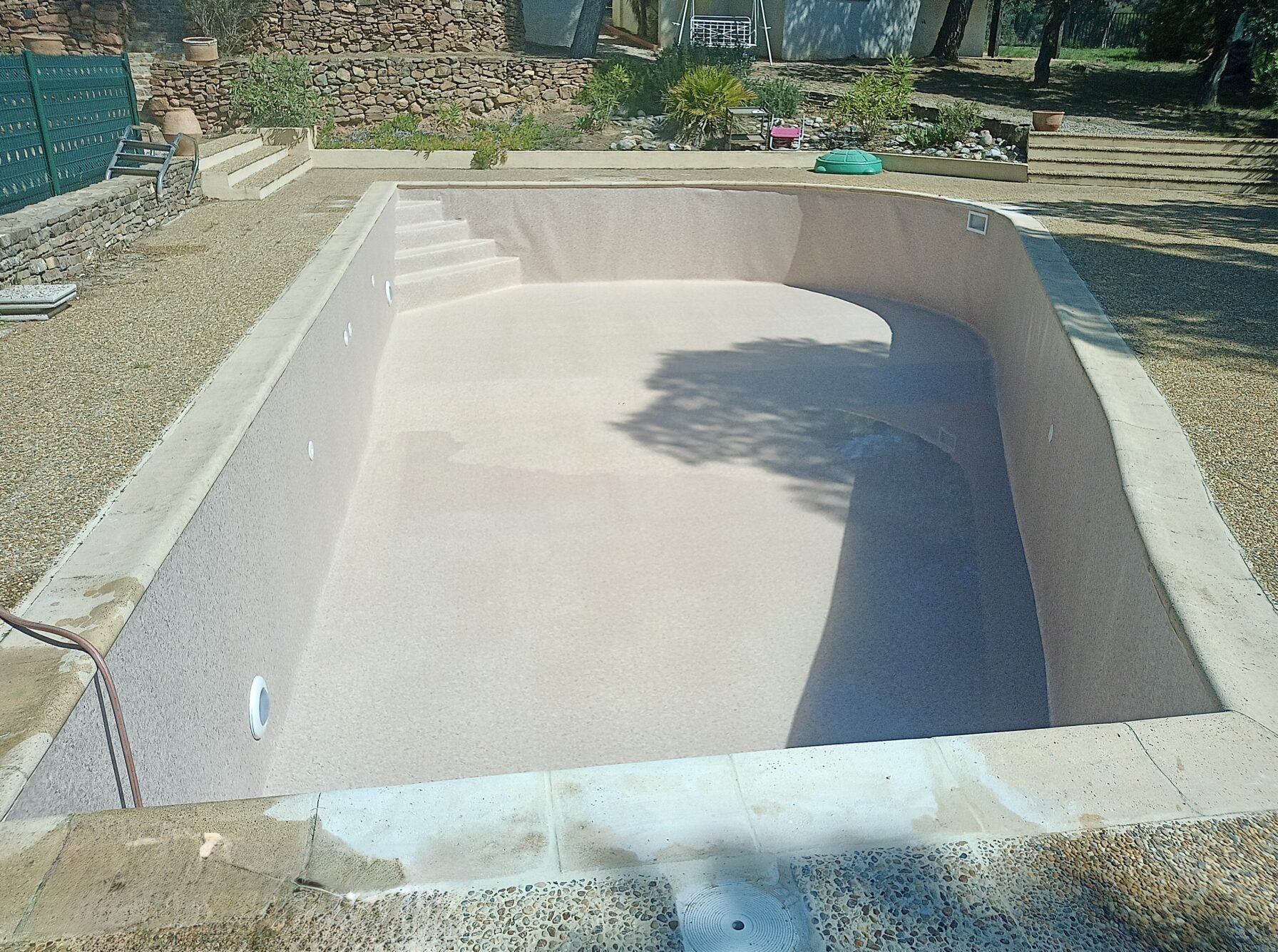Piscine vide à revêtement beige clair avec margelles en pierre et plage en galets dans un jardin paysager.