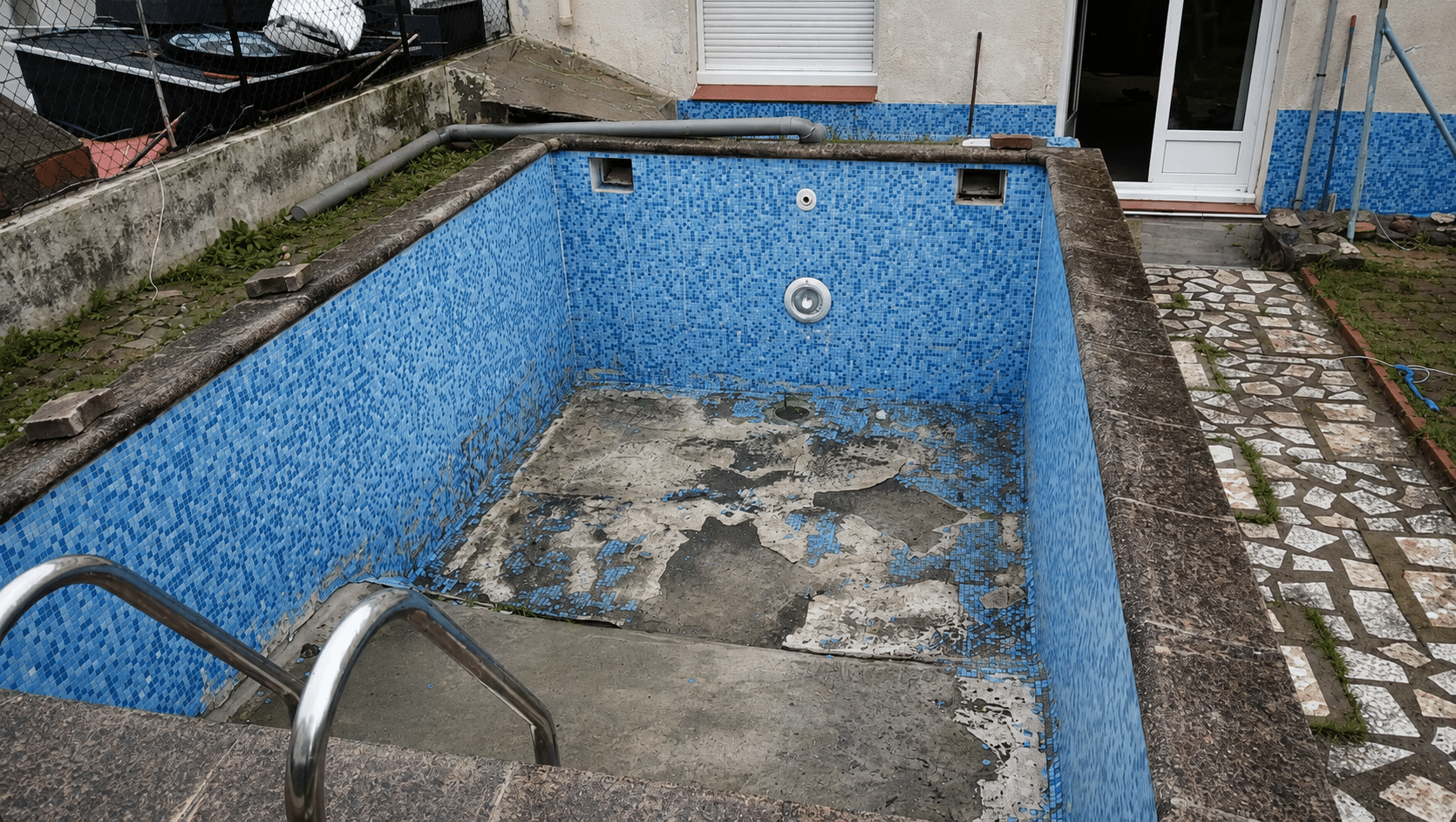 Piscine vide en rénovation avec mosaïque bleue abîmée et fond en béton apparent dans un jardin.