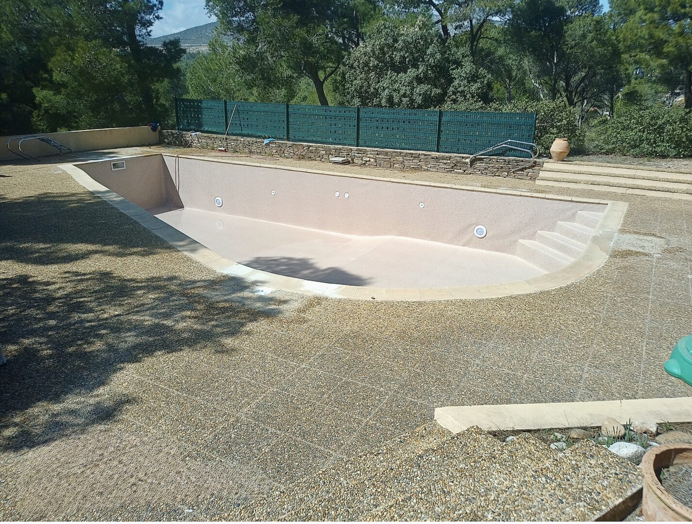 Piscine creusée vide avec terrasse en gravier et clôture de jardin sous un soleil d'été.