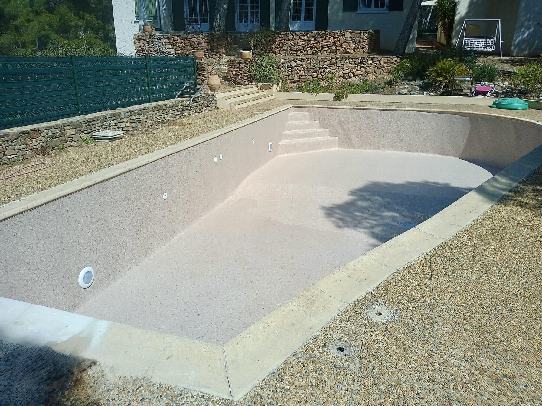 Piscine vide avec revêtement rose sable, escaliers et plage en gravier devant un jardin paysager avec mur en pierre.
