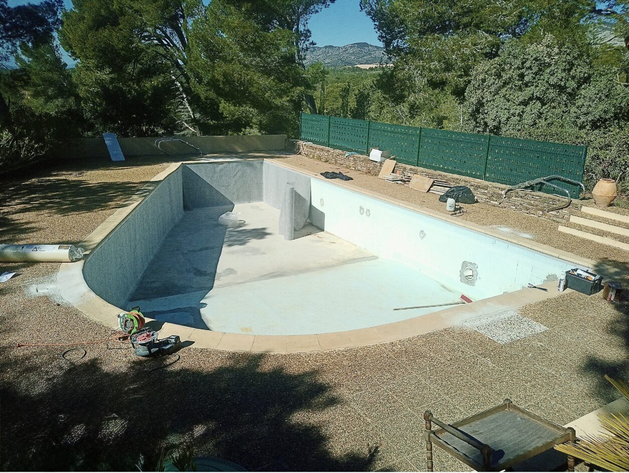 Rénovation d'une piscine vide entourée de gravier, d'arbres et d'une clôture verte avec outils de travaux.