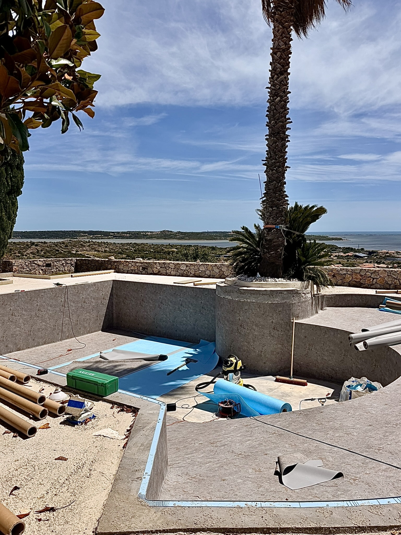 Pose de liner bleu dans une piscine en rénovation avec vue panoramique sur la mer et palmier sous un ciel azur.