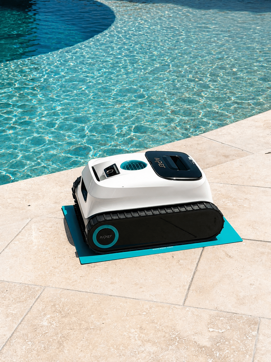 Robot nettoyeur de piscine Aiper blanc et noir sur un tapis turquoise au bord d'une piscine ensoleillée.