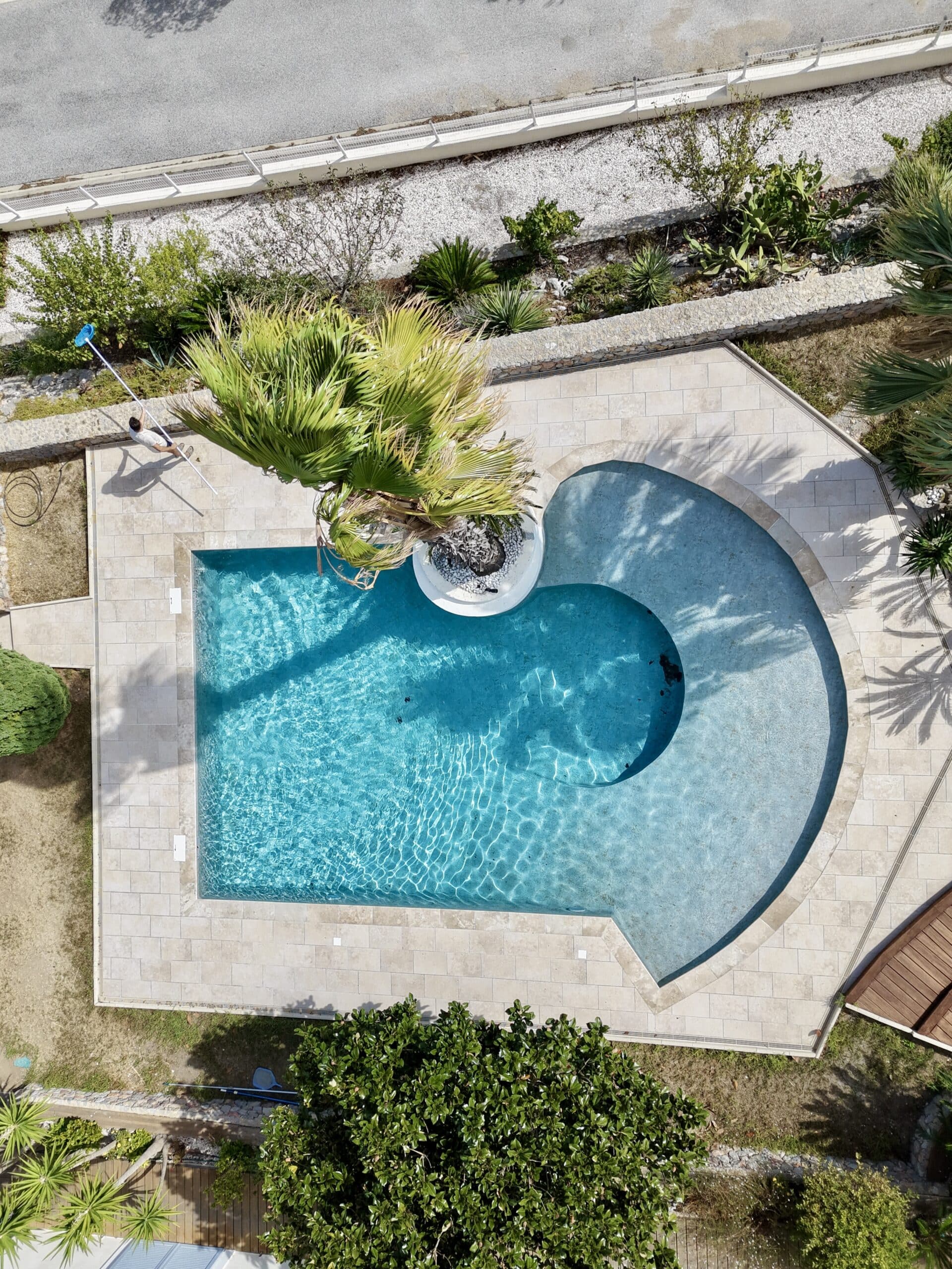 Vue aérienne d'une piscine bleue avec un palmier et une personne passant l'épuisette sur la terrasse en dalles.