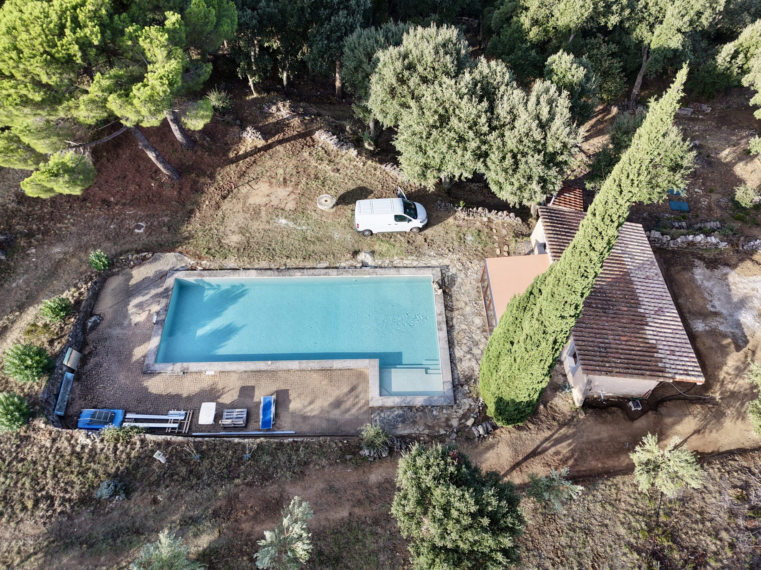 Vue aérienne d'une piscine azur avec terrasse, cabanon provençal et van blanc dans un jardin boisé paisible.